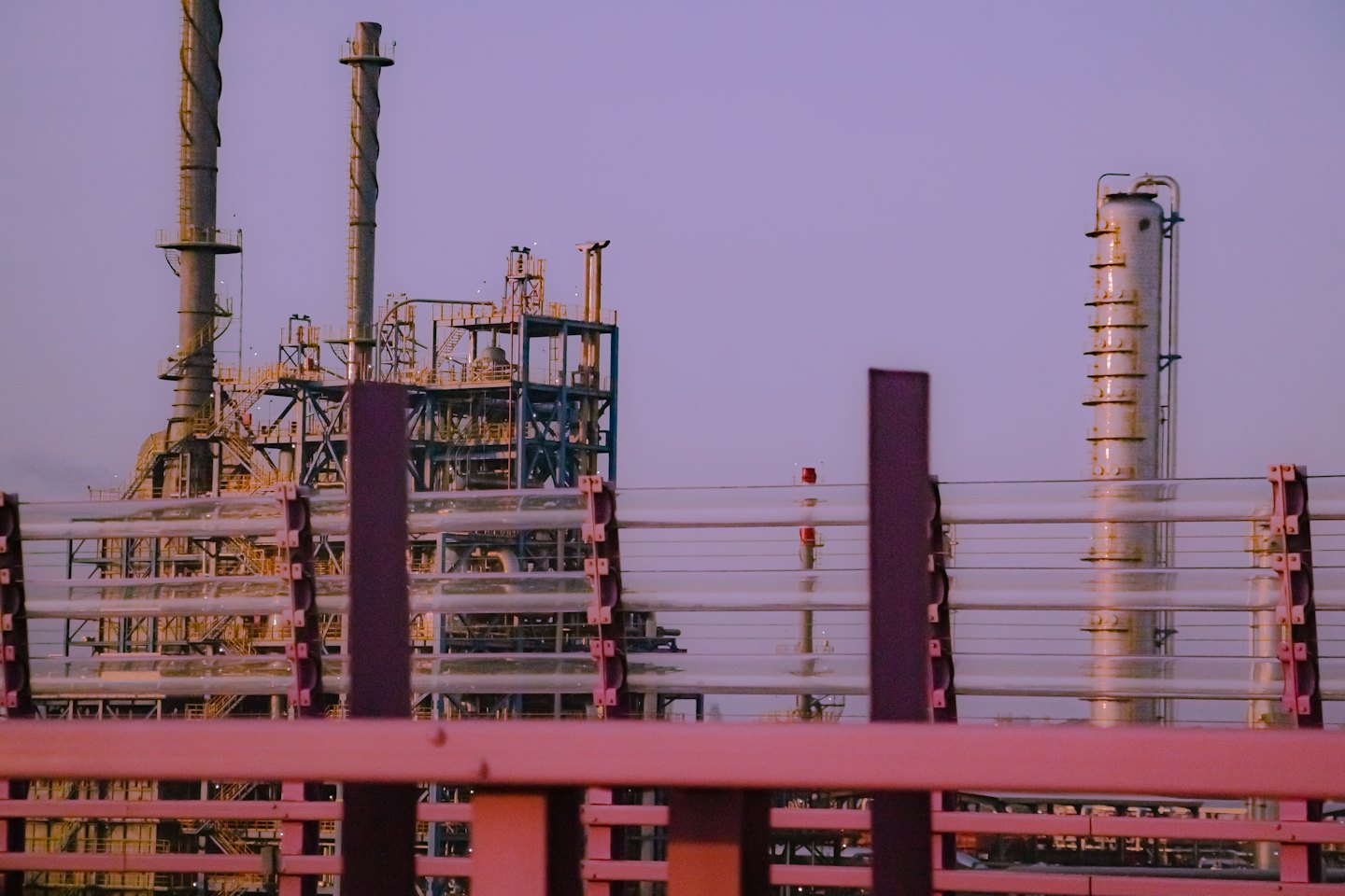 Impianto industriale con ciminiere e tubi dietro una barriera metallica.